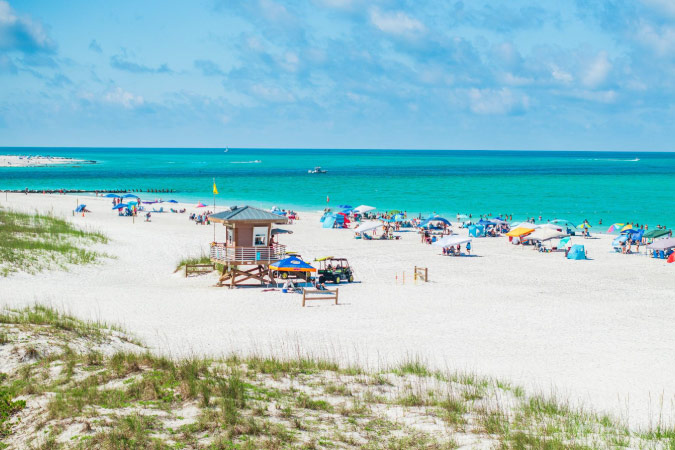 A sunny and beautiful beach in Bradenton, Florida, featuring a quaint lifeguard stand, many colorful umbrellas, white sands, and turquoise waters