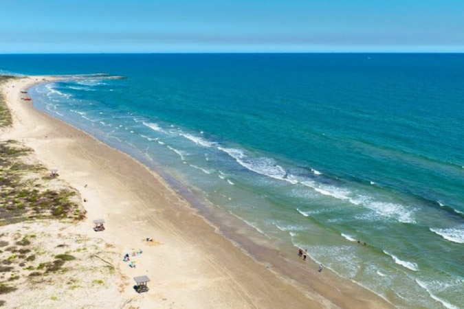 Aerial view of Padre Island National Seashore in Corpus Christi, Texas, on a sunny day, featuring vibrant blue-green waters and an expansive beach.
