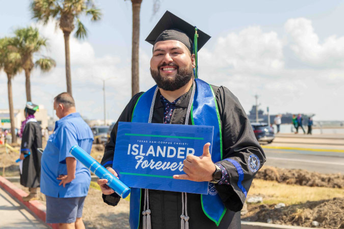 A graduate of Texas A&M University in Corpus Christi is dressed in robe and cap, holding his degree in one hand and throwing a “shaka” sign with his other hand. He’s holding a sign that reads “Islander Forever.”