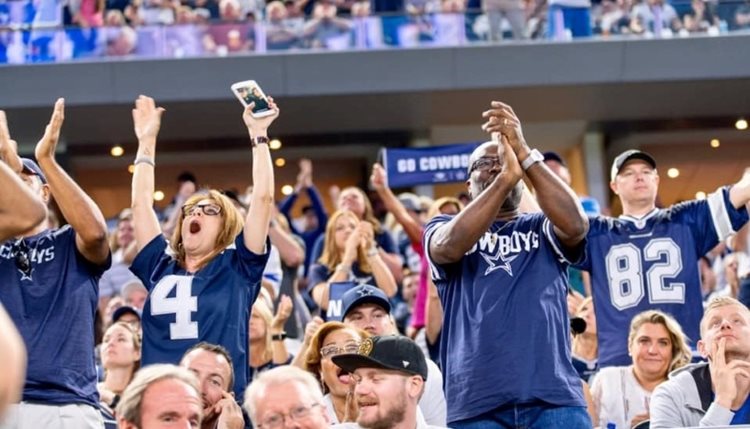 A group of Dallas Cowboys fans are decked out in team jerseys while cheering at a game in AT&T Stadium