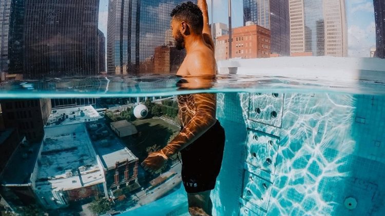 A man is looking down on a Dallas city street as he cools off in a unique, glass-sided, rooftop pool.