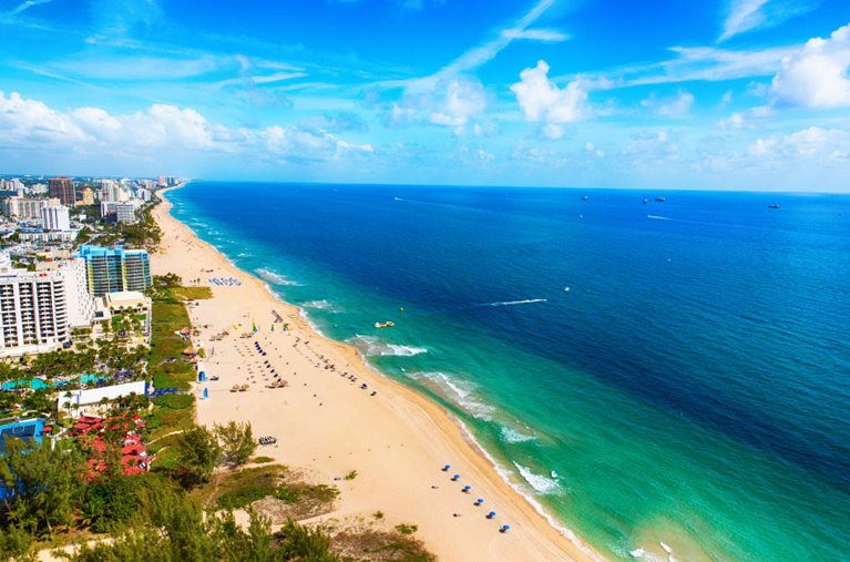 Aerial view of Fort Lauderdale Beach, Florida, showing cream-colored sands lined with rows of blue beach chairs and umbrellas.
