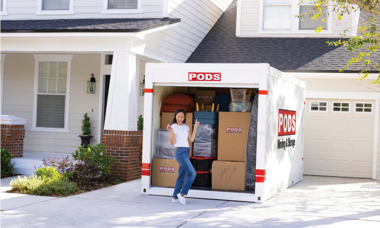 A woman is holding a key in one hand and pointing behind her with the other as she poses in front of her fully loaded ӰPro portable moving container in the driveway of her home. 