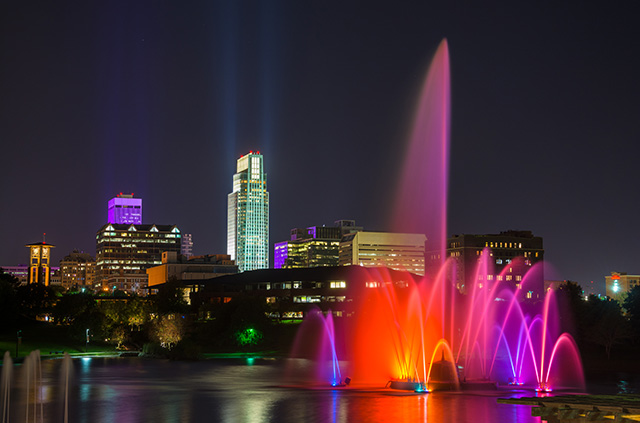 The Omaha skyline at night, featuring illuminated city buildings and a fountain in the foreground, lit by red and purple lights.