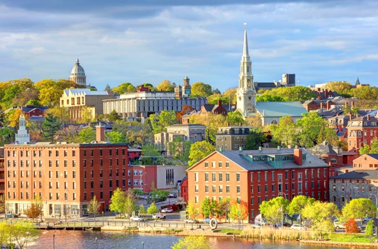 View of Providence, Rhode Island, from across the river. It’s a lovely spring day. The buildings look bright in the sunlight, and the trees are lush with green foliage.