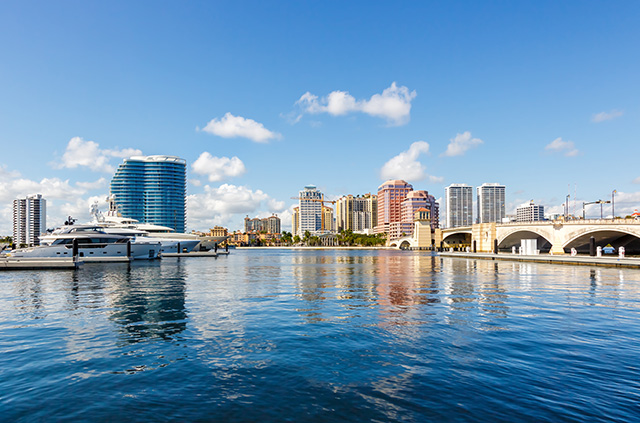 The skyline of West Palm Beach, Florida, during the day. The multi-colored high-rise buildings in the background are reflected on the water in the foreground.