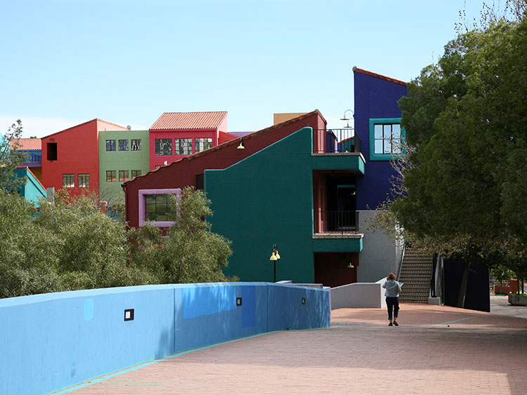 A woman walks across a bridge toward La Placita, a colorful multistory office complex. The buildings are varying shades of red, blue, and green.