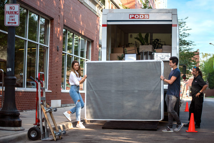 A man and woman are lifting a mattress onto a truck lift so they can load it into a PODS portable moving container, which is still on the bed of the truck.