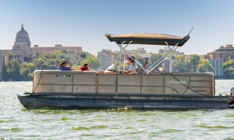 A family is enjoying an afternoon boating on a lake by Madison, Wisconsin. The Downtown buildings are visible in the distance under a clear blue sky.