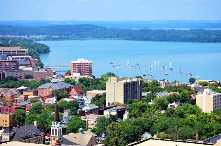 Aerial view of Downtown Madison, Wisconsin, and the surrounding forests and lakes. There are many sailboats out on the water, and the mature trees in the downtown area are lush with green leaves.
