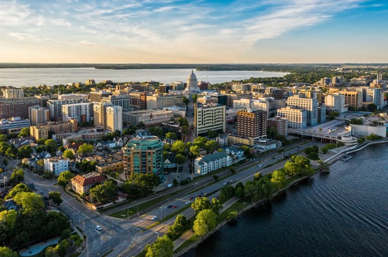 A sunset aerial view of the downtown area of Madison, Wisconsin. The sky is transitioning from blue to peach, and wispy clouds are stretching from the distance. The fading light is reflecting off downtown buildings, and both Lake Mendota and Lake Monona are visible in the picture.