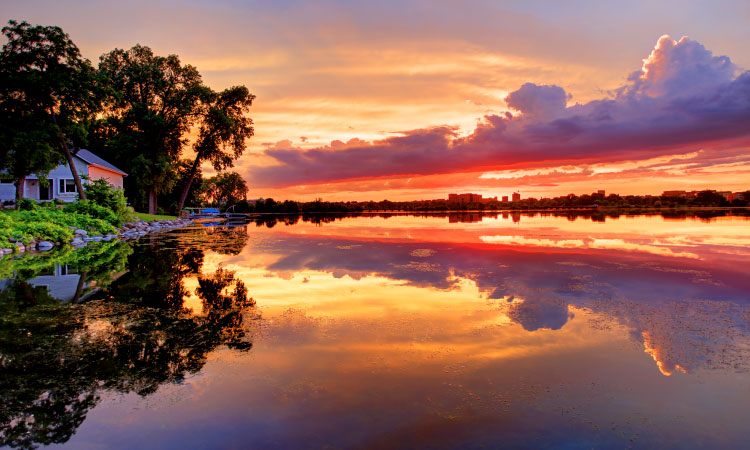 Lake Monona in Madison, Wisconsin, during a stunning sunset. There is a lovely single-family home right on the banks of the lake, and in the distance, under a gorgeous sky, you can see the silhouette of the city of Madison.