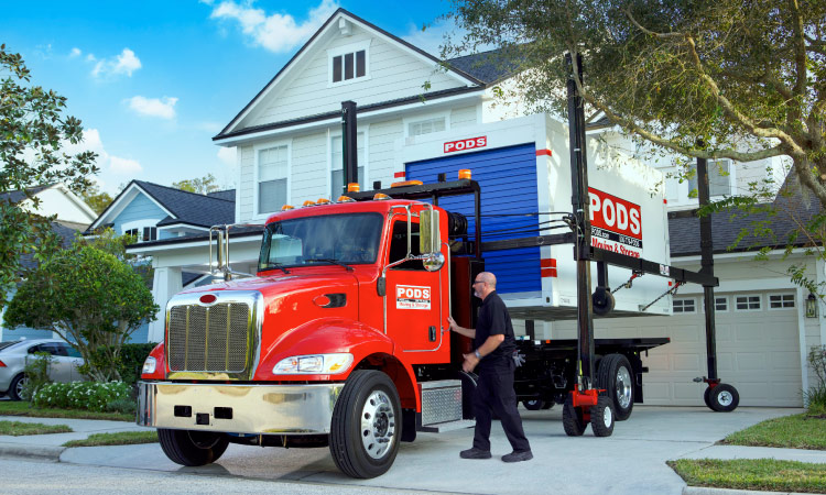 A PODS driver is about to climb into the cab of his signature red PODS truck, which is parked in a residential driveway. The PODS container on the back of the truck has been hoisted up by PODZILLA, and the driver is preparing to move the truck out from under PODZILLA so he can position the container in the driveway for easy loading.