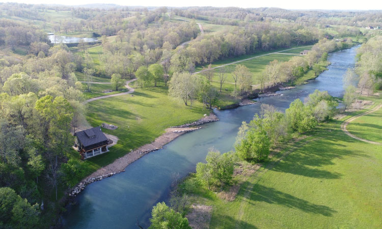 An aerial view of the autumnal beauty along Spring River in Mammoth Spring, Arkansas. The river's sandy shore is gracefully bordered by trees, most of which have shed their leaves for the season. Amidst this picturesque landscape, a charming residential home stands alongside the tranquil riverbank. The scene showcases the serene essence of autumn as it unfolds along the scenic Spring River.