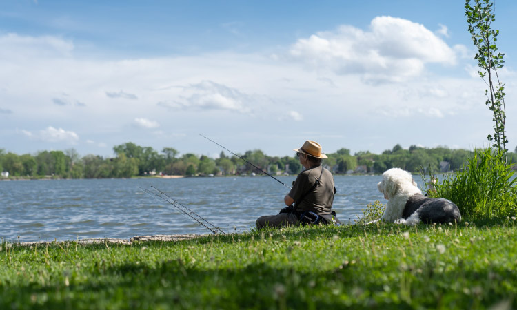 A man is relaxing with his dog by a lake in Madison, Wisconsin. It’s a sunny summer day and the man is wearing sunglasses and a straw hat. He’s sitting on a lush carpet of grass on the banks of the lake, holding a fishing rod as he waits for a fish to take the bait. There are two other fishing rods stuck in the sand nearby.