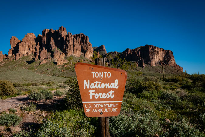 A freestanding sign reads “Tonto National Forest” and is posted in front of the Superstition Mountains in Arizona.