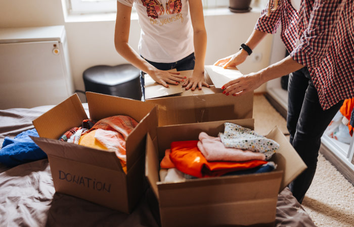 A mother and daughter are closing one of three cardboard boxes filled with clothing donations. They’re decluttering and minimizing their belongings to save on interstate moving costs.