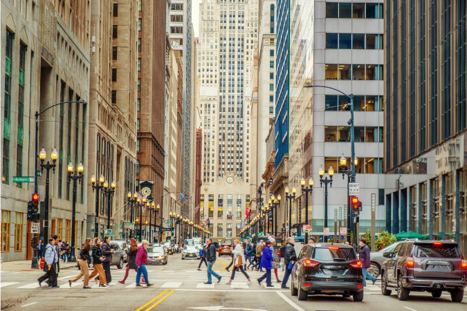 Dozens of pedestrians are crossing a main thoroughfare between massive skyscrapers in Chicago, Illinois.