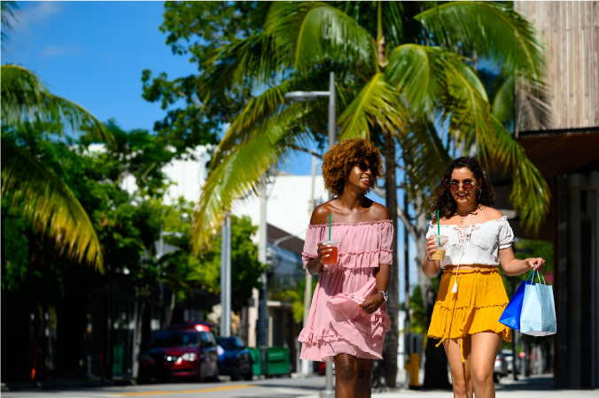 Two women in colorful summer outfits are holding to-go coffees and paper shopping bags as they stroll along the streets of Miami.