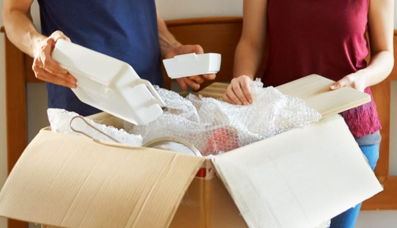 A man and a woman are packing dishes with bubble cushioning roll into a moving box for their PCS move.