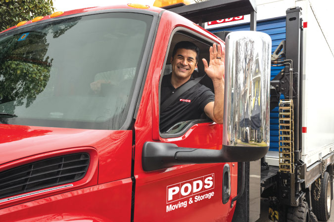 A PODS truck driver is smiling and waving from the cab of his truck as he prepares to transport a PODS portable moving container for a long-distance move.