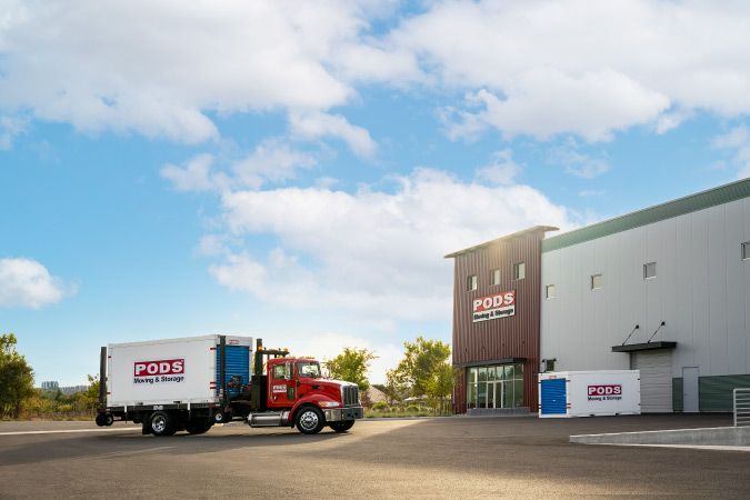 Exterior view of a PODS storage facility with a portable storage container parked outside and a PODS truck arriving with another container.