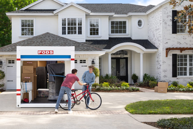 A father hands his teenage son a bicycle as they unload their PODS container at their new home.