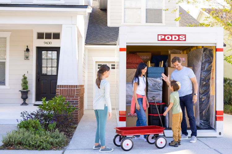 A family of four celebrates loading up their PODS portable moving container. It’s in their driveway and is neatly packed with furniture and moving boxes. 