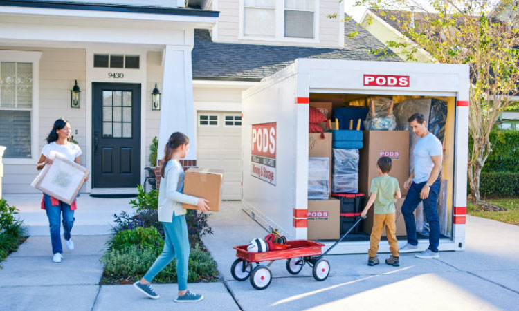 A family of four is working together to load the last of their things into a PODS portable moving container in their driveway. The mother and daughter are carrying framed pictures and a moving box from inside the house. The boy has a red wagon with some sports equipment, and the father is standing by the door of the container, ready to load it with these final items.