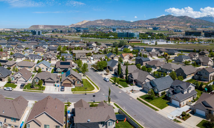 An aerial view of a large residential area in Salt Lake City. The neighborhood consists of mostly two-story homes with green lawns and the occasional mature tree sprouting up from a side yard. A range of mountains is rising up in the distance.