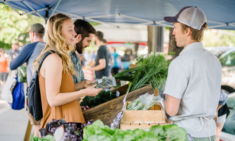 A young woman is shopping at a produce stand at the Downtown Farmers Market in Salt Lake City, Utah. She’s smiling as she examines a bag of herbs and speaks with the farmer behind the stand. 