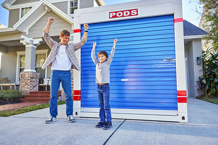 A dad and son celebrate loading the last of their belongings in their PODS portable moving and storage container. They are holding their arms in the air as if cheering, with the container and their two-story gray house behind them. 