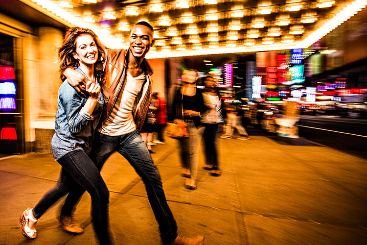 A young couple walks out of a Broadway theater, smiling arm-in-arm