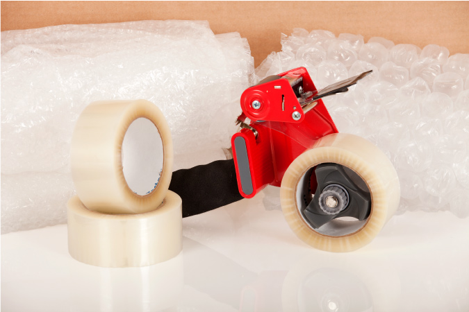 Packing supplies, including a packing tape dispenser with two extra rolls of packing tape beside it and bubble cushioning roll behind it. 