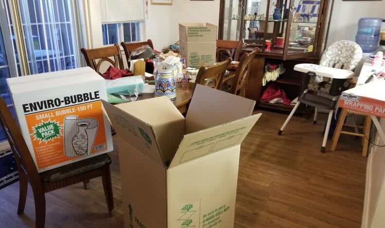 A dining room filled with furniture and several moving boxes and supplies.