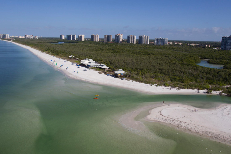 Aerial view of Pelican Bay in Naples, Florida. White sand beaches line the coast, and there’s a thick wooded area between the sands and the big city in the distance. 