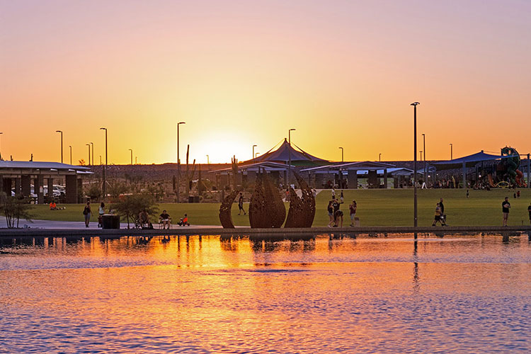 The town of Peoria in Arizona. There is a lake at sunset with people spread out in a green park.  