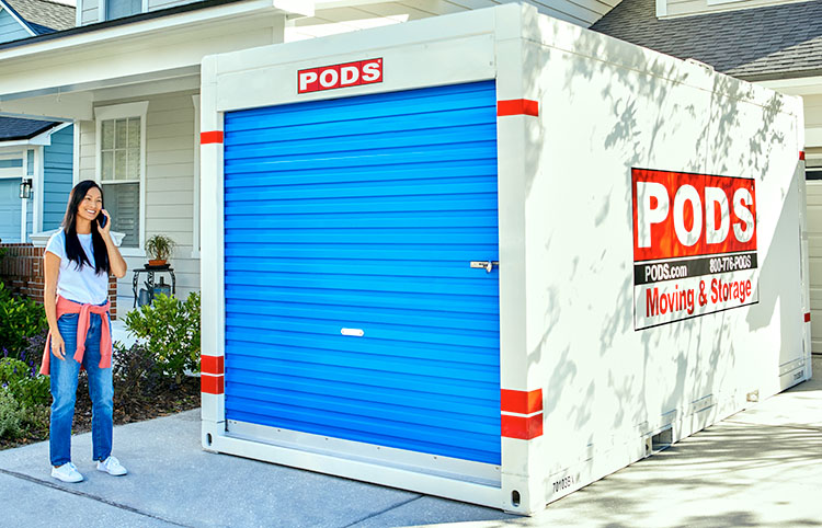 A woman stands in front of a PODS container in her driveway, calling PODS to schedule her move to the Phoenix suburbs. 