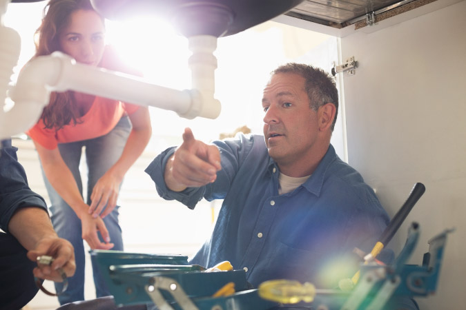 A plumber is leaning under a kitchen sink, explaining something to the homeowners during their kitchen remodel