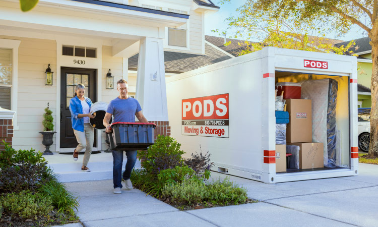 A mature couple is carrying things out of their house to load into the PODS portable moving container in their driveway. The container is nearly full and contains a mattress, moving boxes, and some furniture.