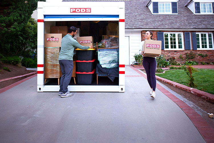 A woman carries a PODS-branded cardboard box out to a loaded PODS container with its door open in her driveway. A man is stacking totes and boxes in the container.