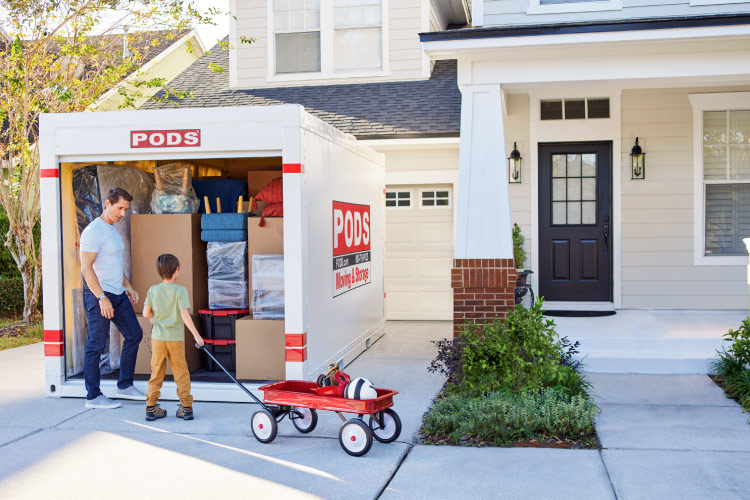 A father and son stand at the entrance to their PODS portable moving container in the driveway of their new home in Providence, Rhode Island. The boy is holding the handle to a red wagon filled with a sports helmet and ball. 