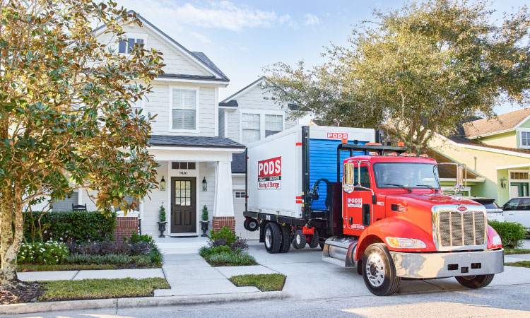 A PODS truck with a portable moving and storage container on the back has just reversed into a residential driveway to deliver the container. The cab of the truck is painted a vibrant red, and the container is white with a blue door. 