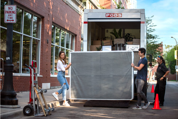 A young couple prepares to lift a mattress into their PODS portable moving container as the PODS driver stands nearby.