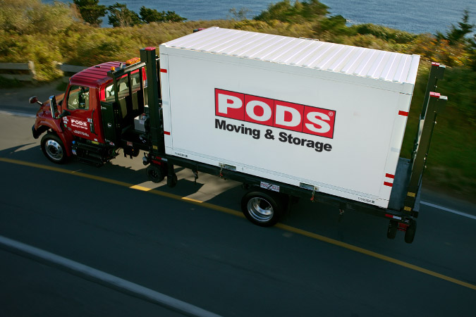 Aerial view of a PODS truck transporting a PODS portable moving container on a coastal road.