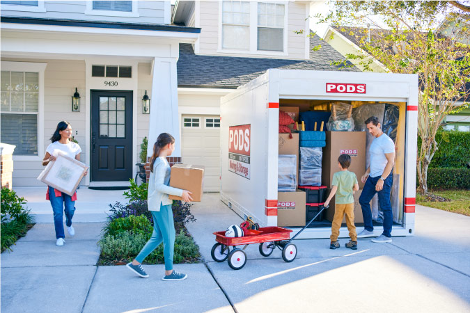 A family of four is carrying boxes and other things out to a PODS portable moving container in their driveway to load it up for their move.