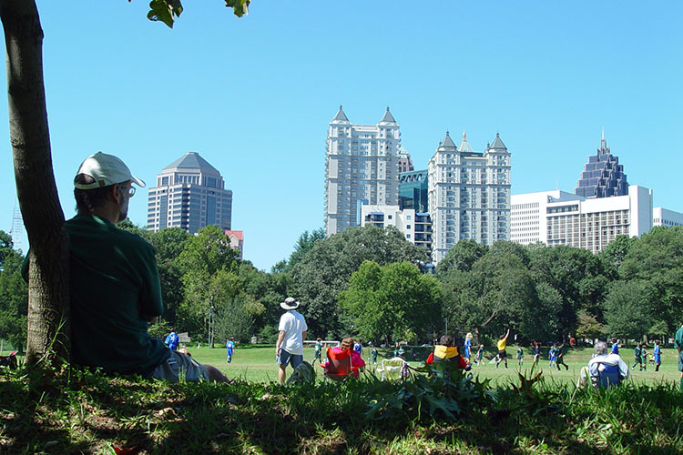 A man in a hat sits under a shady tree, looking out over an Atlanta park. You can see the Atlanta skyline over the trees.