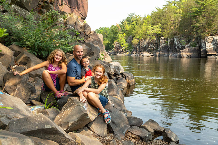 A father and his three daughters sit on the rocks on the riverbank of the St. Croix River