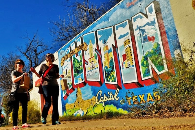 Two women walking in Austin, Texas, on a sunny day. They're passing a colorful mural that reads, "GREETINGS FROM AUSTIN CAPITOL OF TEXAS.