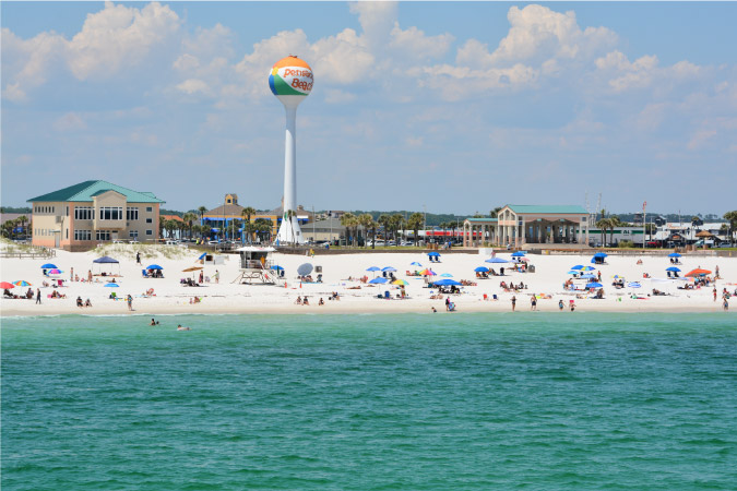 View of Pensacola Beach from out in the water, featuring dozens of beachgoers and a colorful water tower shaped like a beach ball.